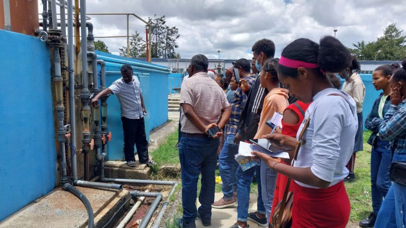 A member of AAWSA staff points at pipes and filters as he explains and demonstrates water treatment equipment to a group of school pupils